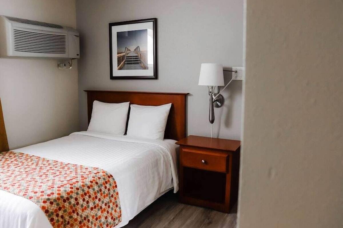 A hotel room with a bed, a nightstand with a lamp, an air conditioner, and a framed picture on the wall.