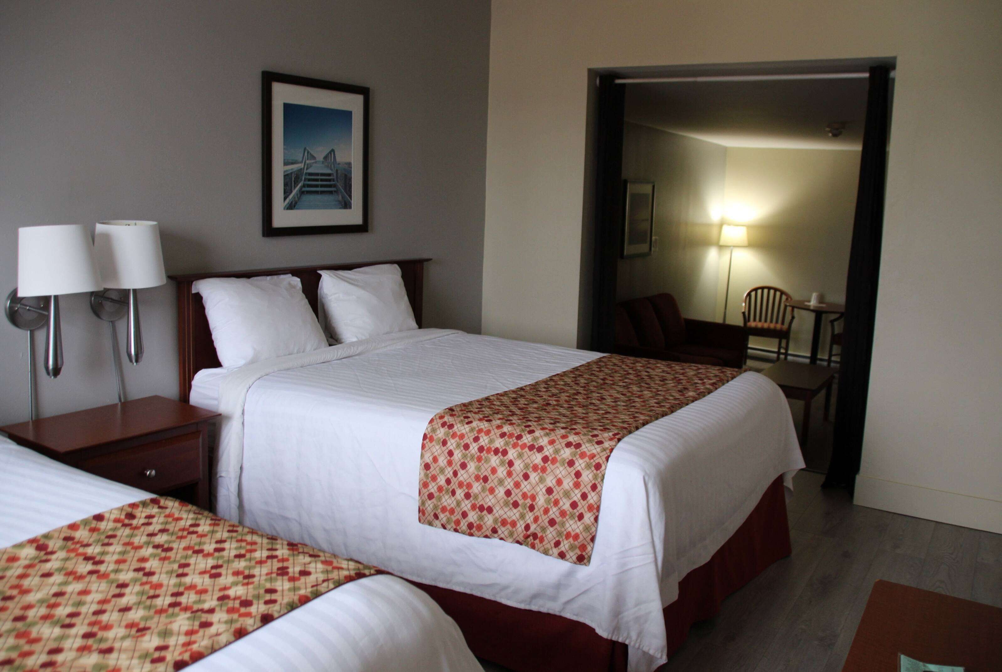 A hotel room with two beds, a nightstand, a lamp, a chair, and a door leading to another room.