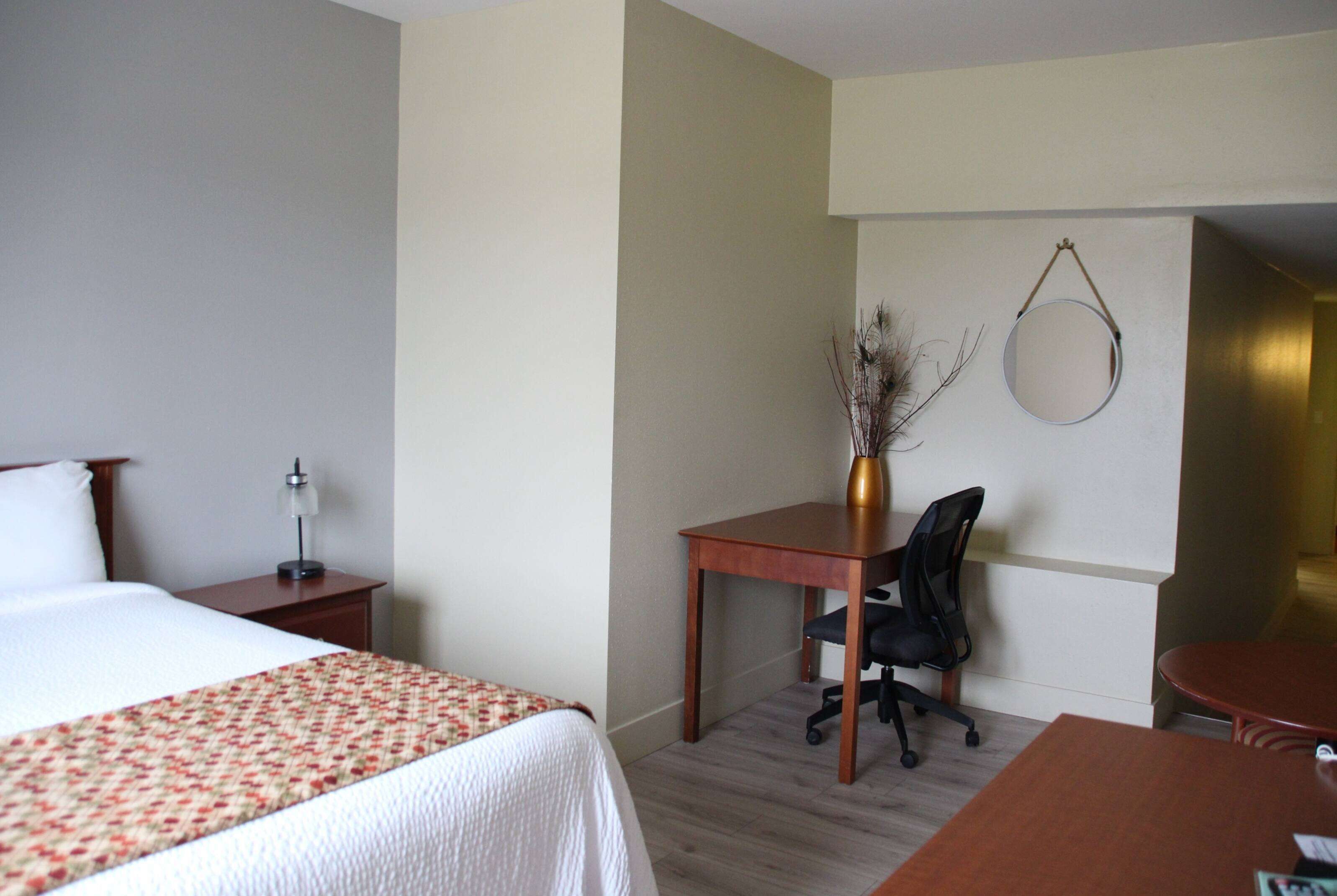 Habitación de hotel con cama, escritorio con silla, una mesita pequeña y un espejo redondo en la pared.
