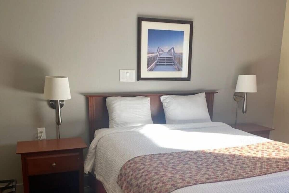 A hotel room with a bed, two bedside tables with lamps, a framed picture on the wall, and a wooden nightstand.