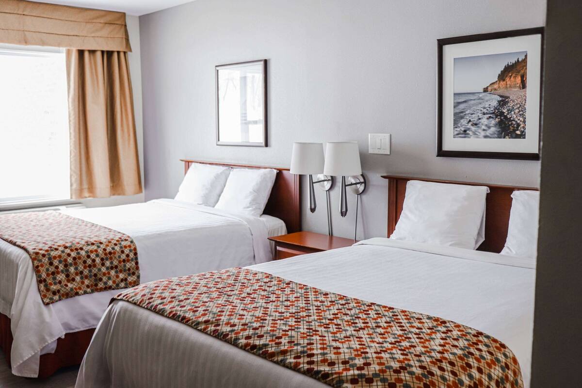 A hotel room with two beds, a bedside table, a lamp, a framed picture on the wall, and a window with curtains.