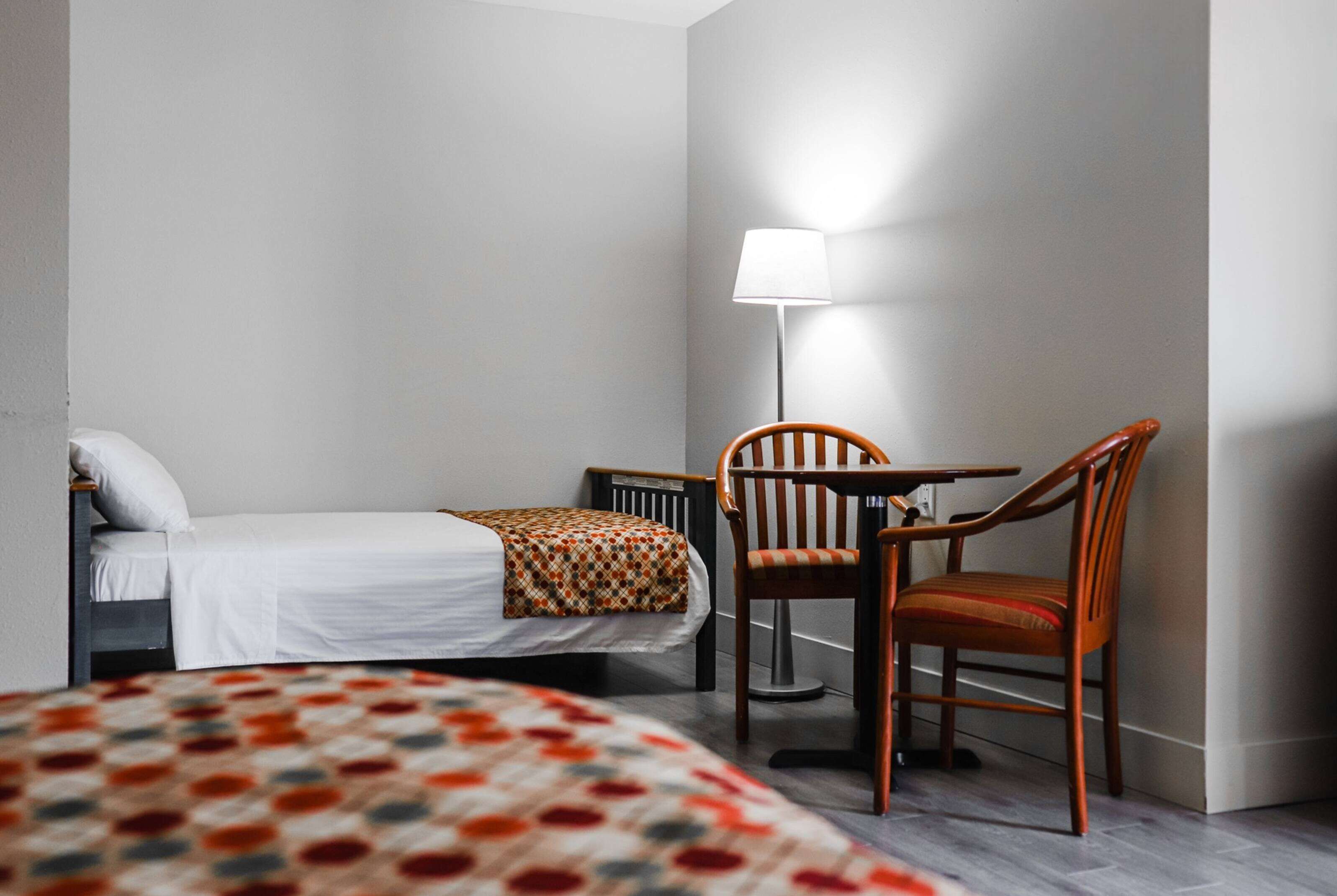 A single bed with a patterned bedspread, a wooden chair, a small table, and a floor lamp in a room with plain walls.
