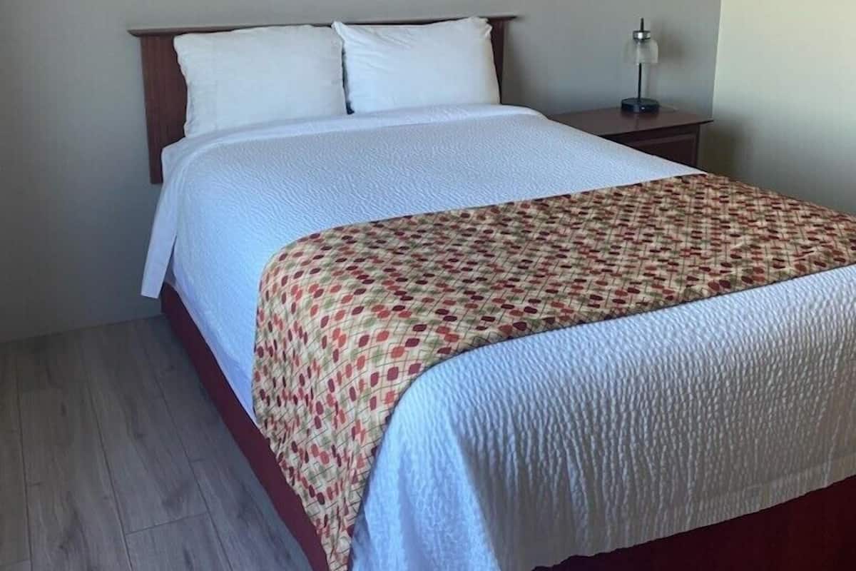 A neatly made bed with a white comforter and a patterned throw blanket, a bedside table with a lamp, and a wooden floor.