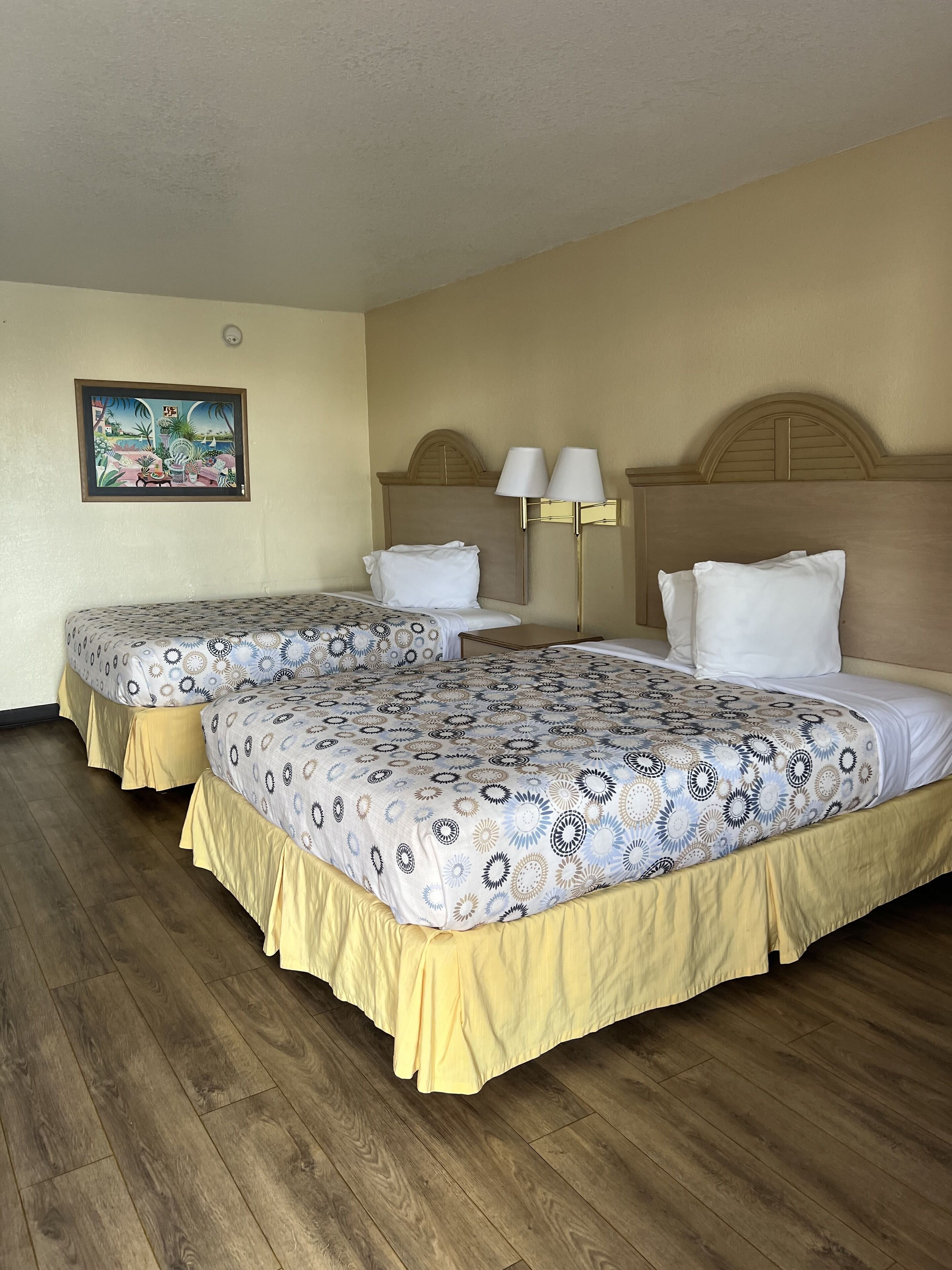 A hotel room with two beds, a painting on the wall, and a nightstand with lamps.