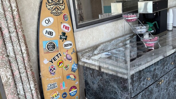 Un área de recepción con una tabla de surf decorada con pegatinas, una planta en maceta con una bandera estadounidense y una barra de mármol.