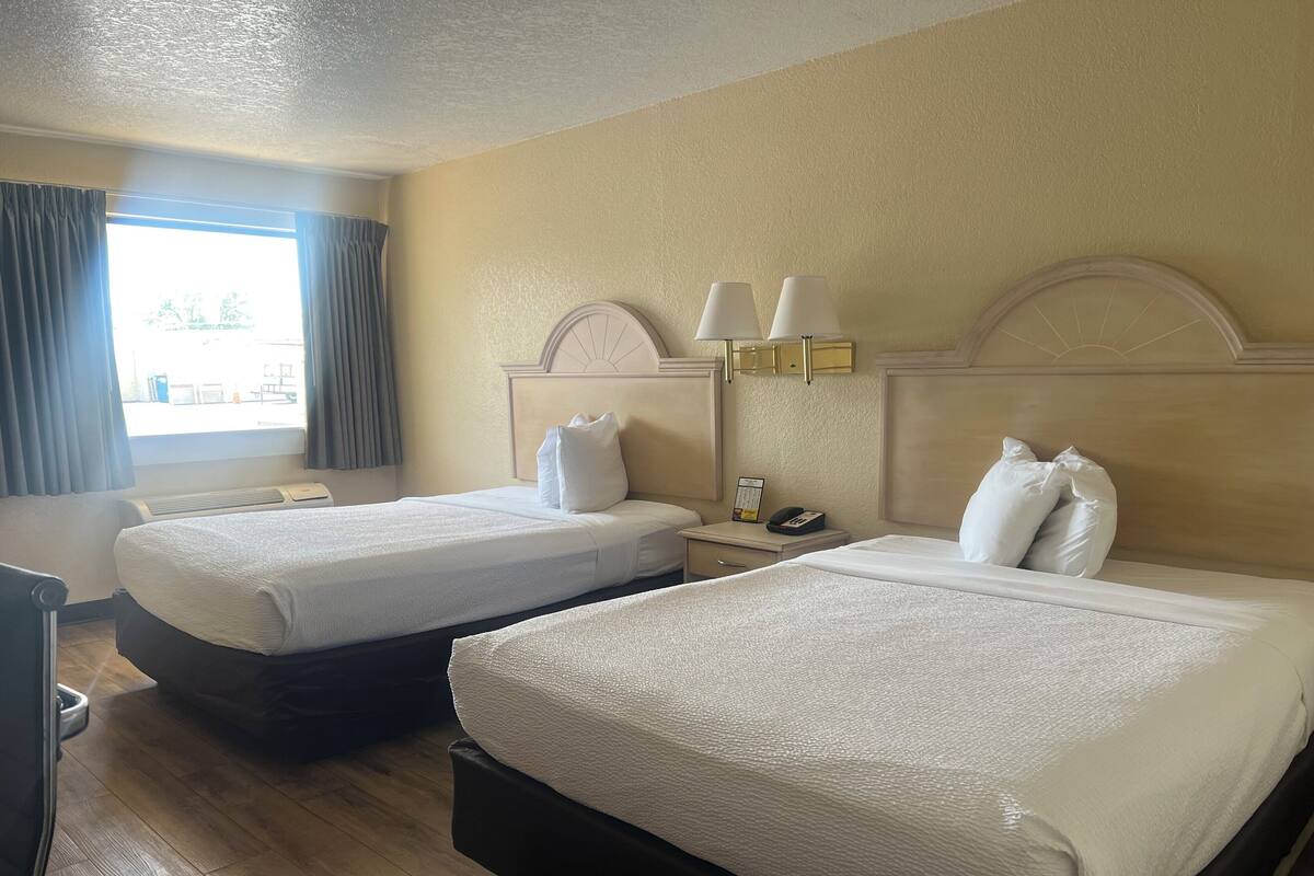 Ein Hotelzimmer mit zwei Betten, einem Nachttisch mit Lampe, einem Fenster mit Vorhängen und einer Wanduhr.