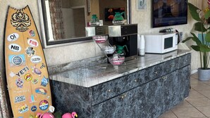 Zona de recepción de hotel con mostrador de mármol, una tabla de surf con pegatinas, una máquina de café y una planta en maceta con la bandera estadounidense.