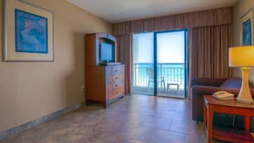 A hotel room with a large sliding glass door to a balcony, a wooden dresser, a desk with a lamp, and a framed abstract painting on the wall.