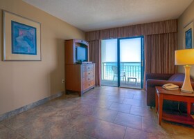 A hotel room with a large sliding glass door to a balcony, a wooden dresser, a desk with a lamp, and a framed abstract painting on the wall.