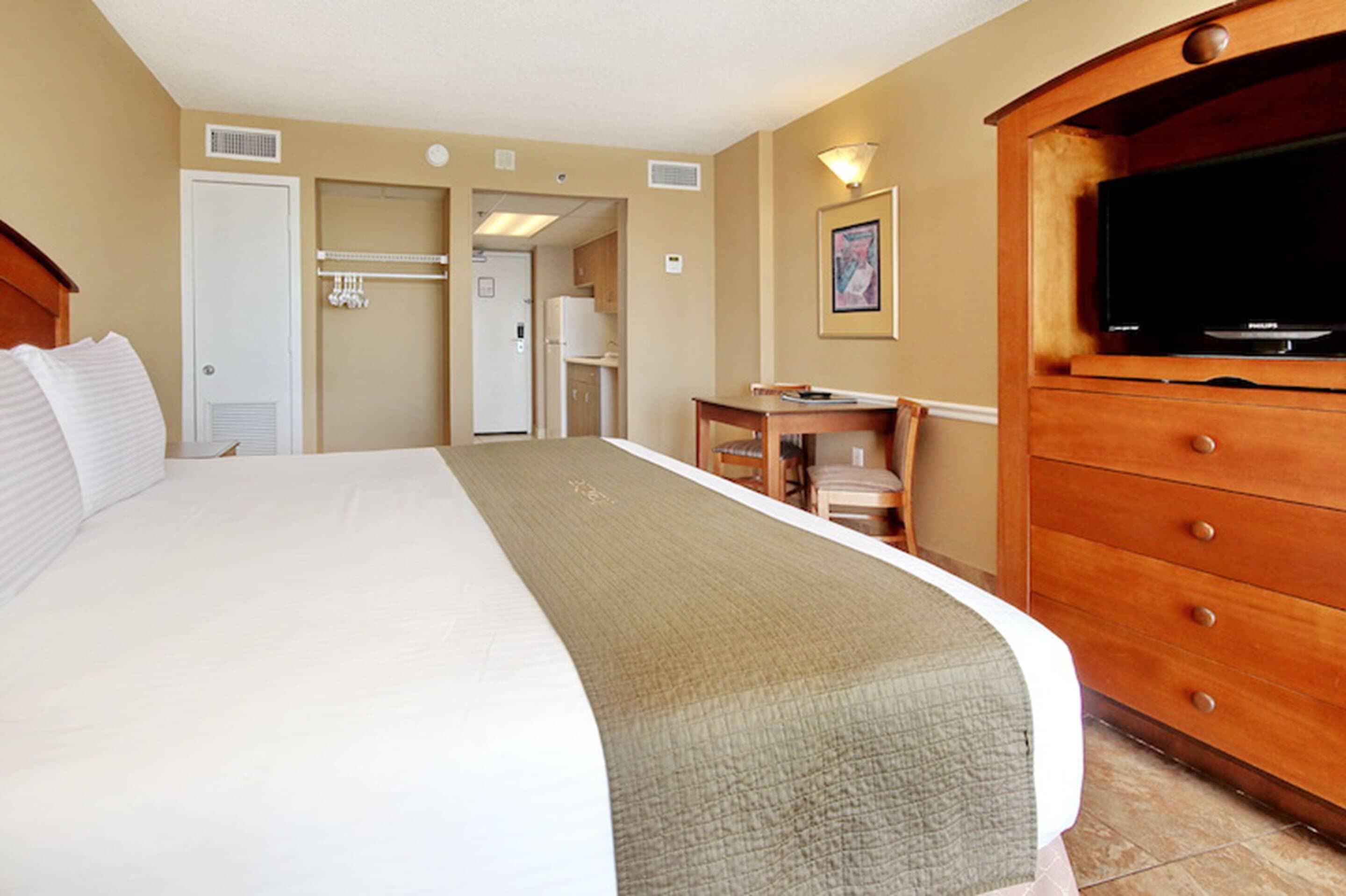 A hotel room with a large bed, a wooden dresser with a television, a desk, and a kitchenette in the background.