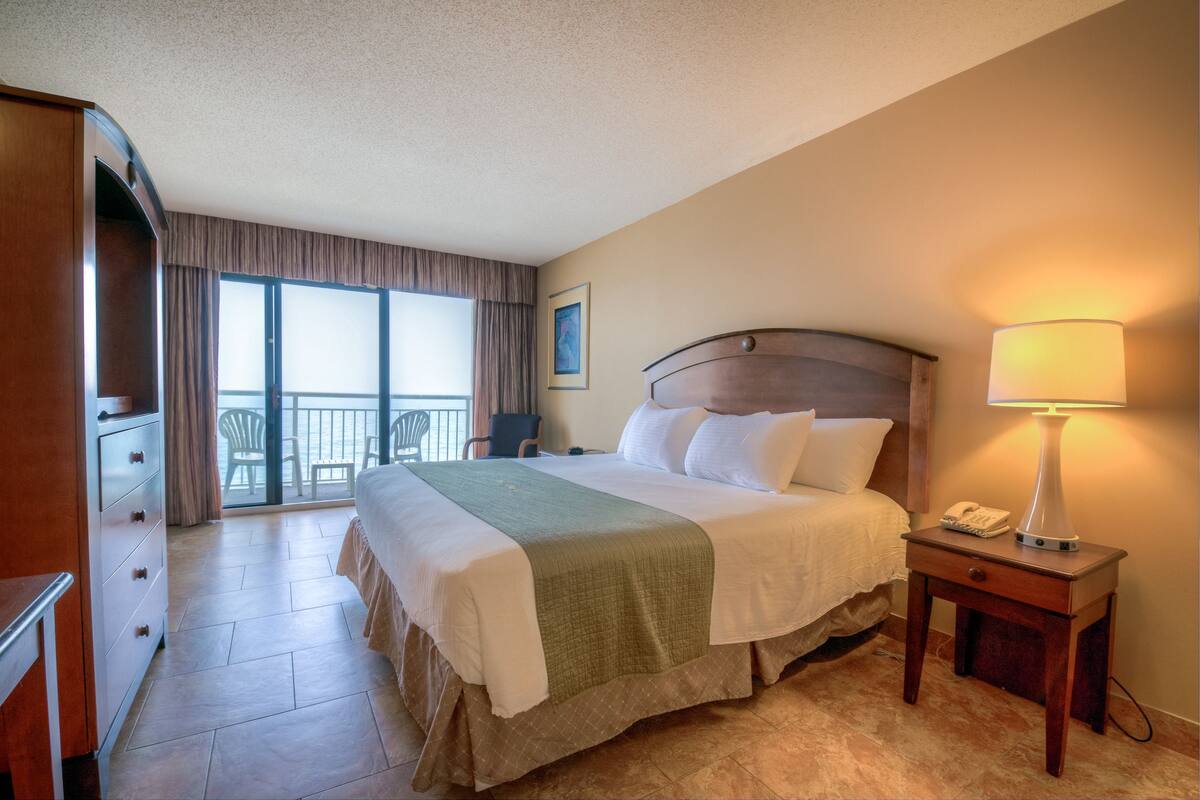 A hotel room with a bed, a nightstand, a lamp, a dresser, and a view of the outside balcony.
