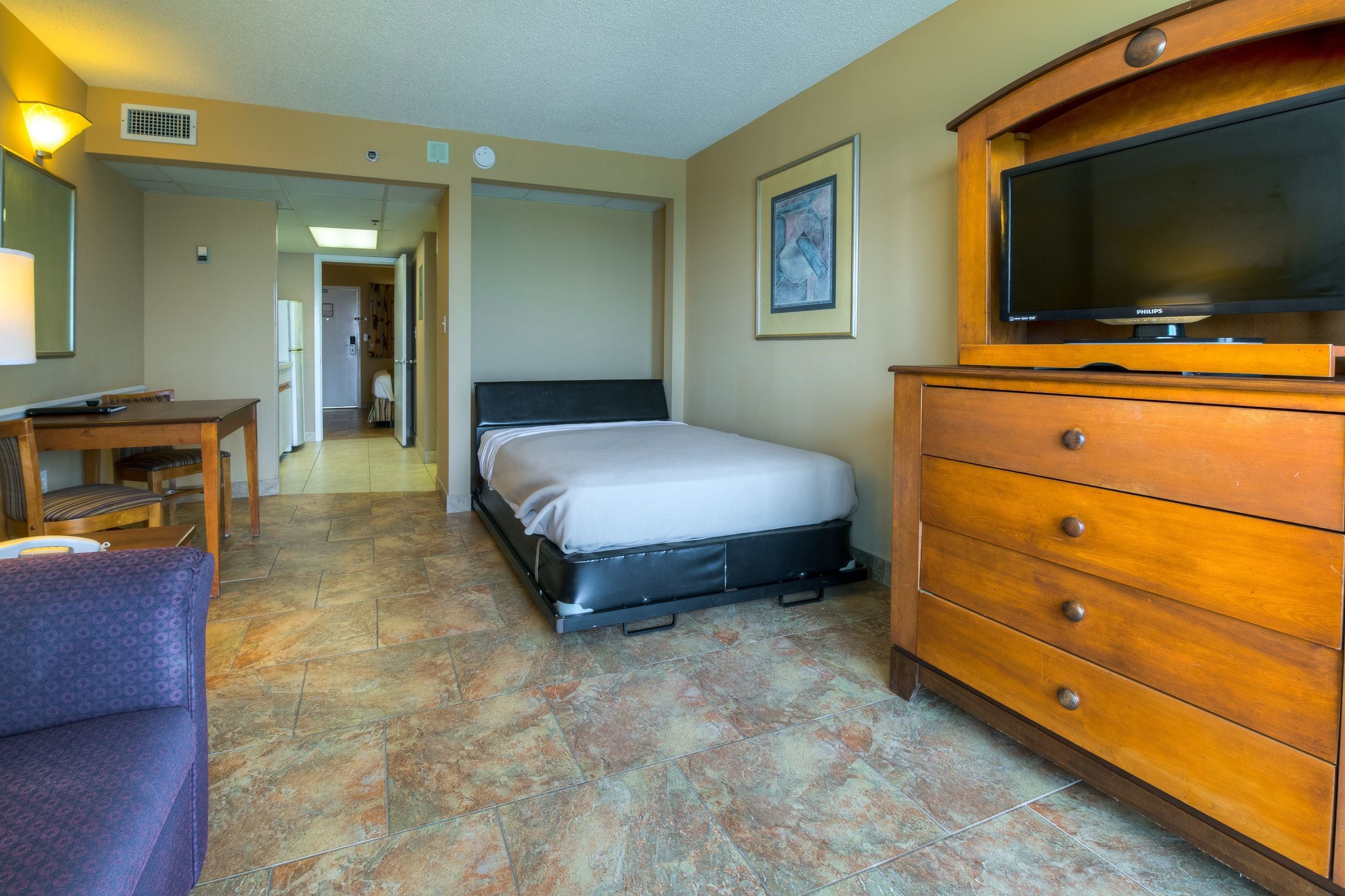 A hotel room with a bed, a wooden dresser, a television, a desk, and a chair.