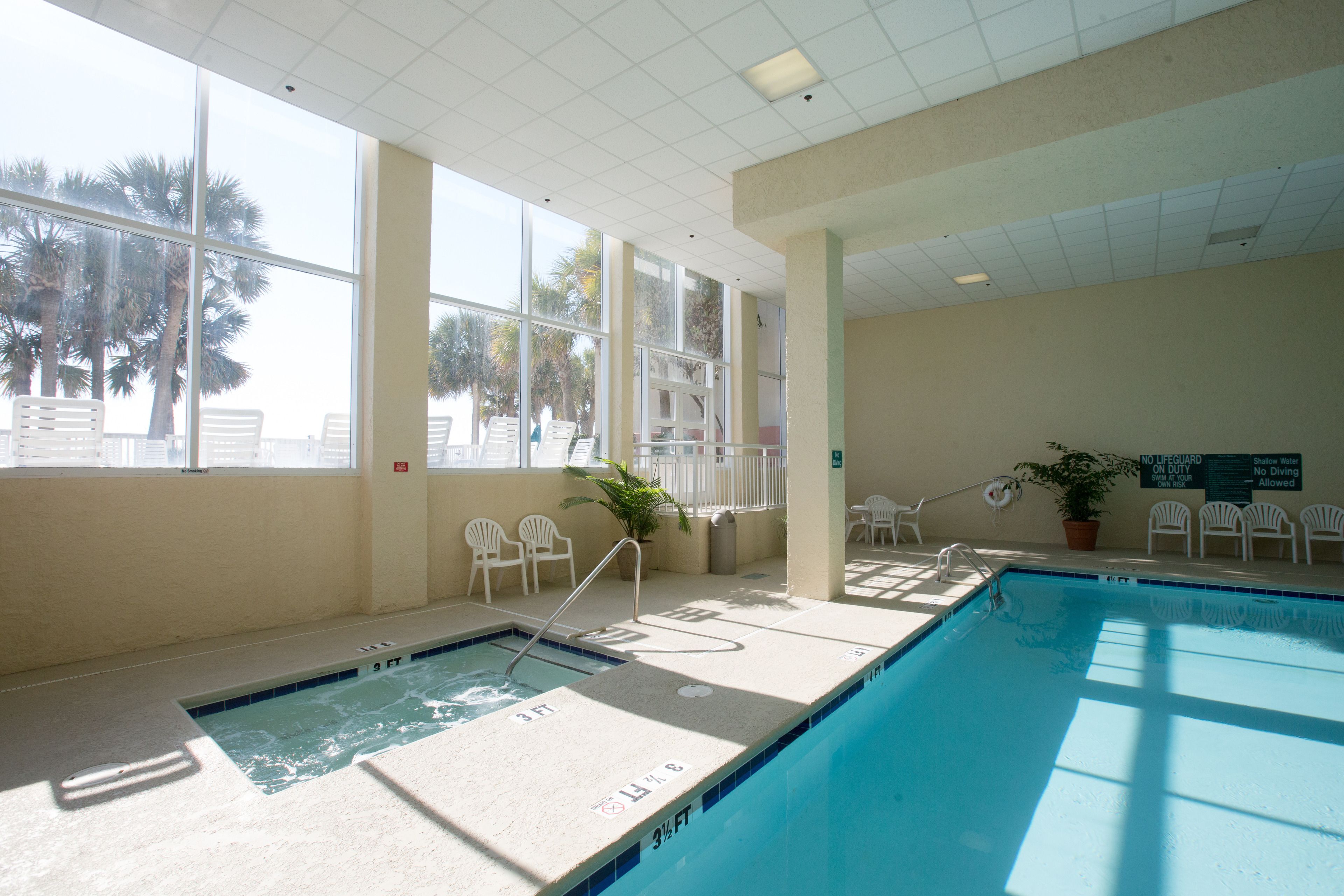 An indoor swimming pool with clear water, surrounded by white chairs and a potted plant.