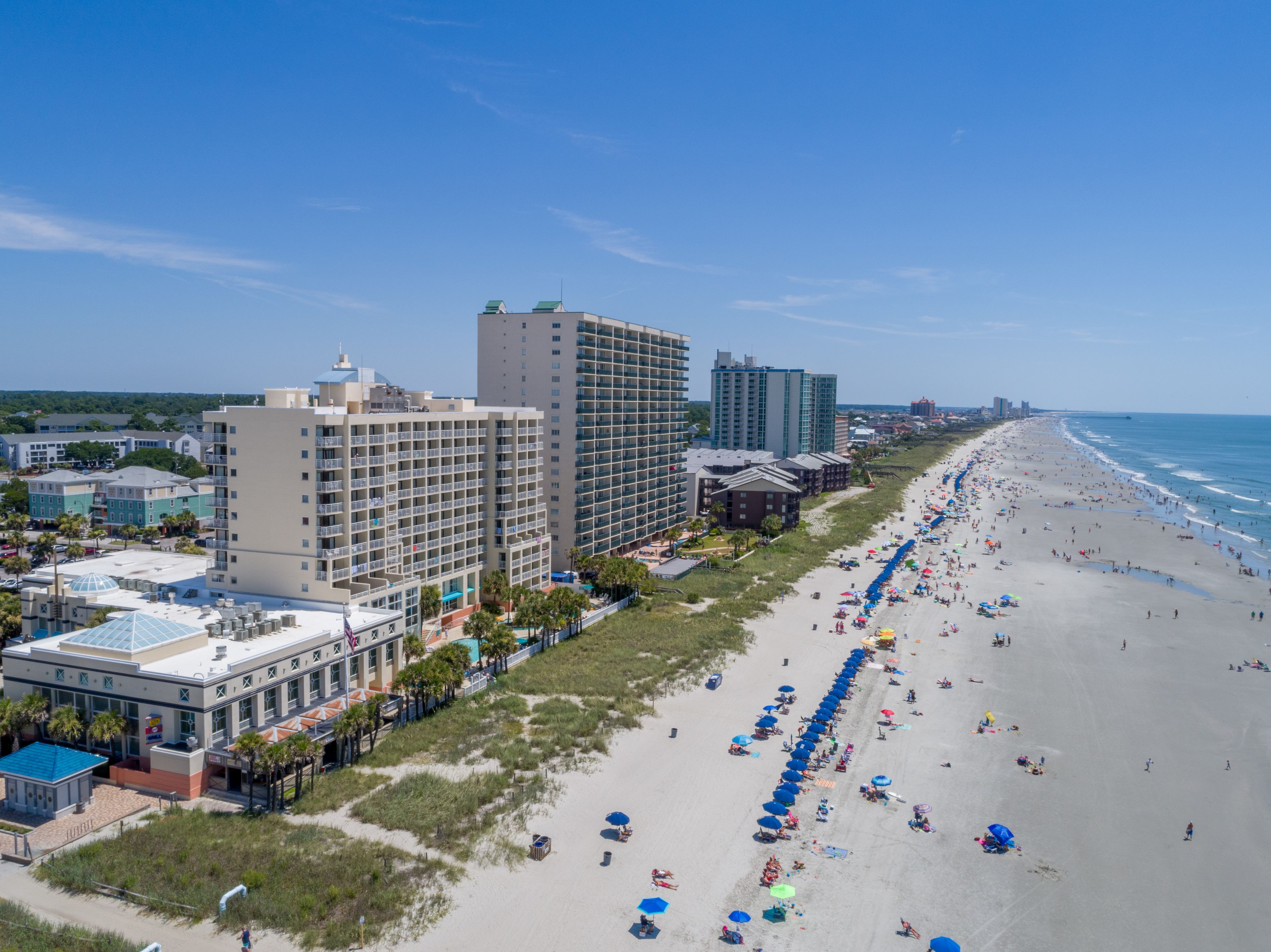 A beachfront hotel with multiple floors, a sandy beach with umbrellas, and a clear view of the ocean.