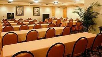 Uma sala de conferências com mesas e cadeiras dispostas em fileiras, uma planta grande e quadros nas paredes.