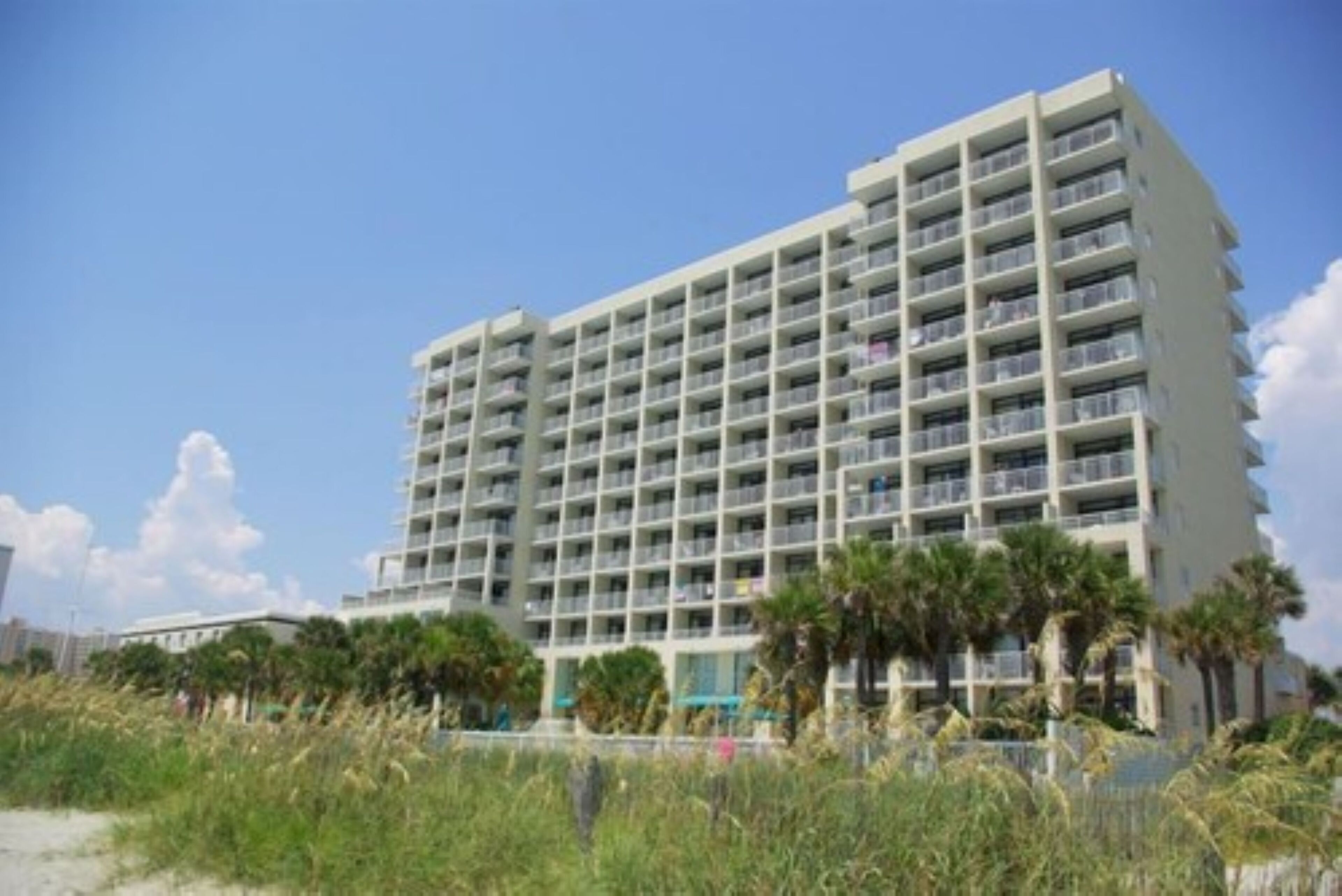 A multi-story hotel with balconies, located near a beach with palm trees.