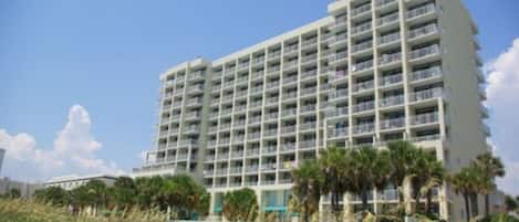 A multi-story hotel with balconies, located near a beach with palm trees.