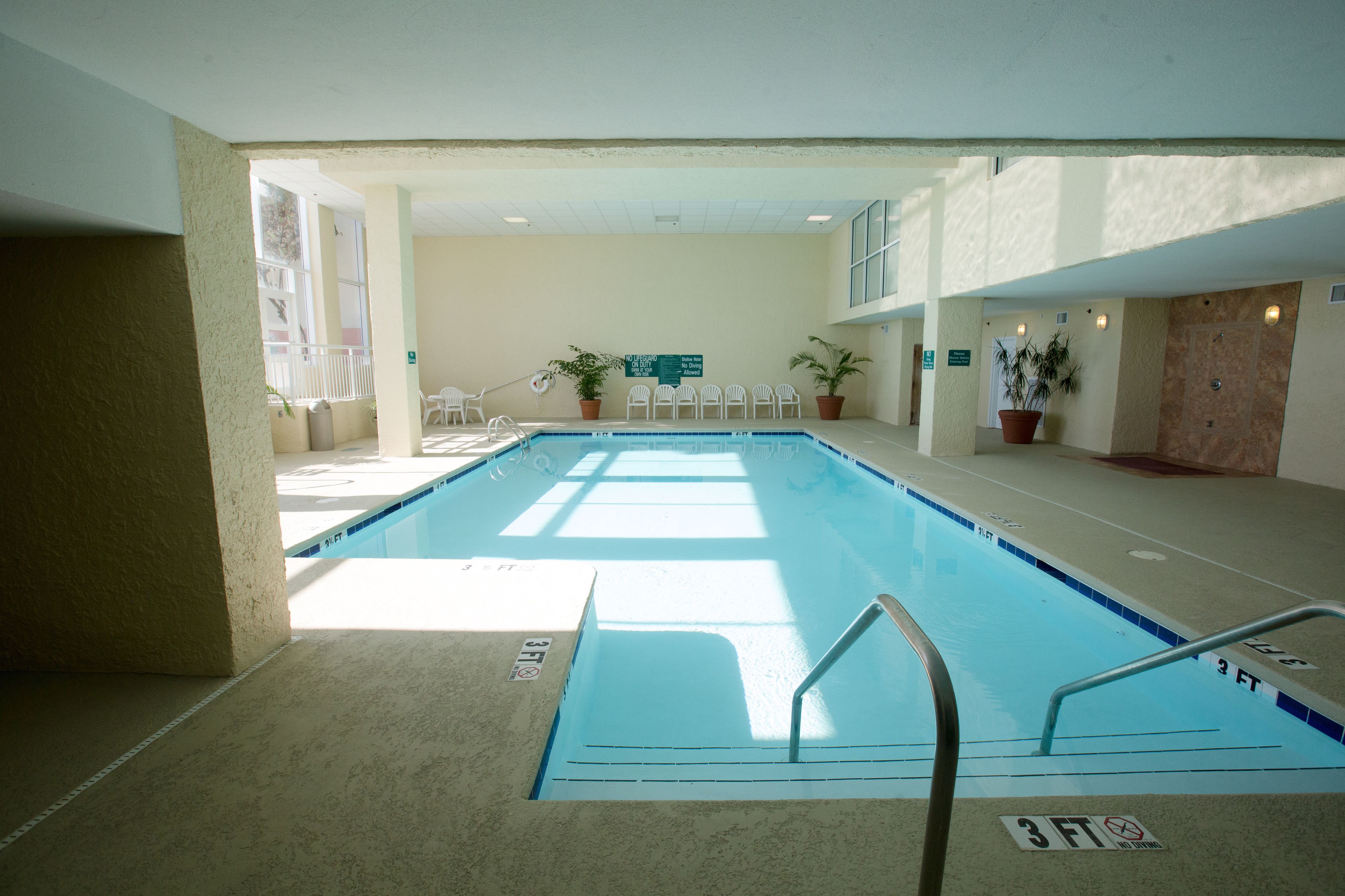 An indoor swimming pool with a 3 FT depth marking, surrounded by potted plants and white chairs.