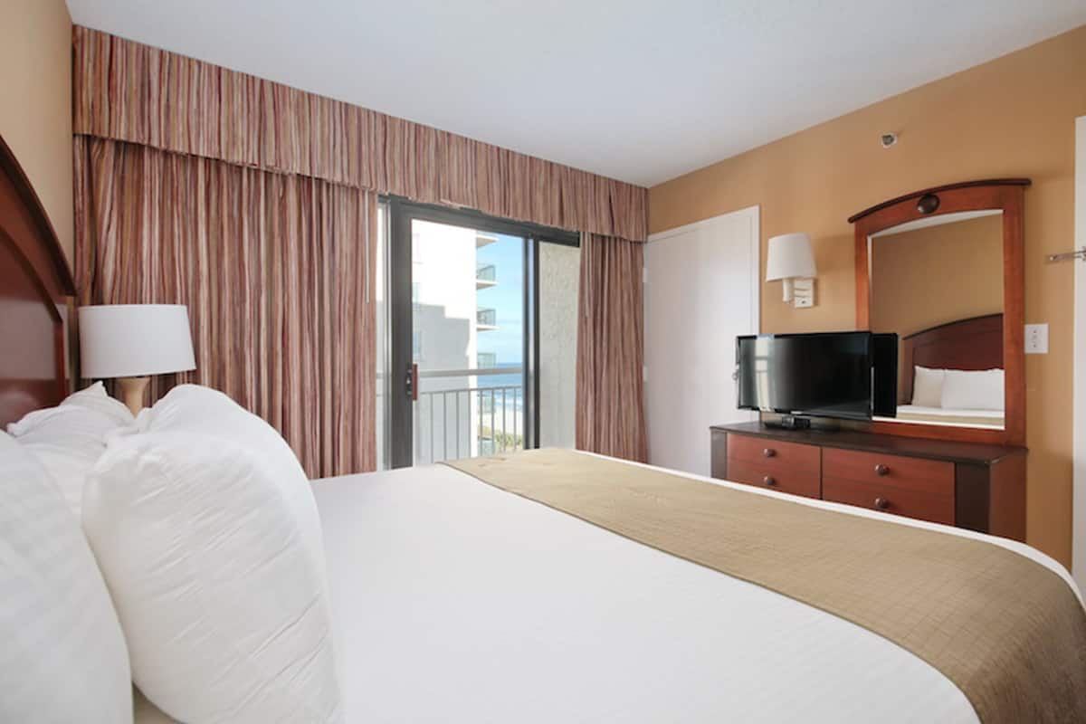 A hotel room with a large bed, a TV on a wooden dresser, a balcony with curtains, and a view of the city.