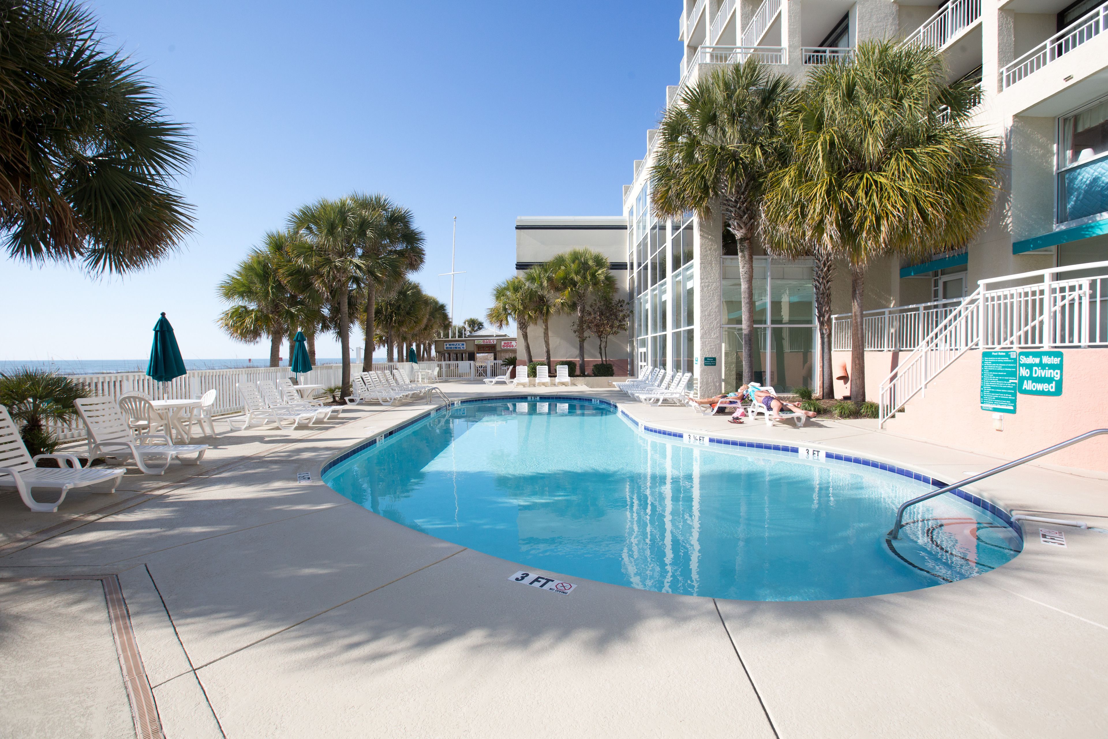 A pool area with lounge chairs and umbrellas, a sign indicating no diving allowed, and a view of the ocean.