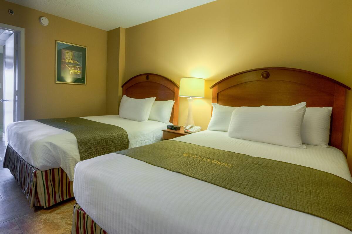 A hotel room with two beds, each with a wooden headboard, white linens, and a green throw blanket. There is a bedside table with a lamp and a phone.