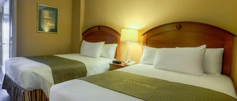 Habitación de hotel con dos camas, cada una con cabecero de madera, ropa blanca y una manta verde. Hay una mesita de noche con una lámpara y un teléfono.
