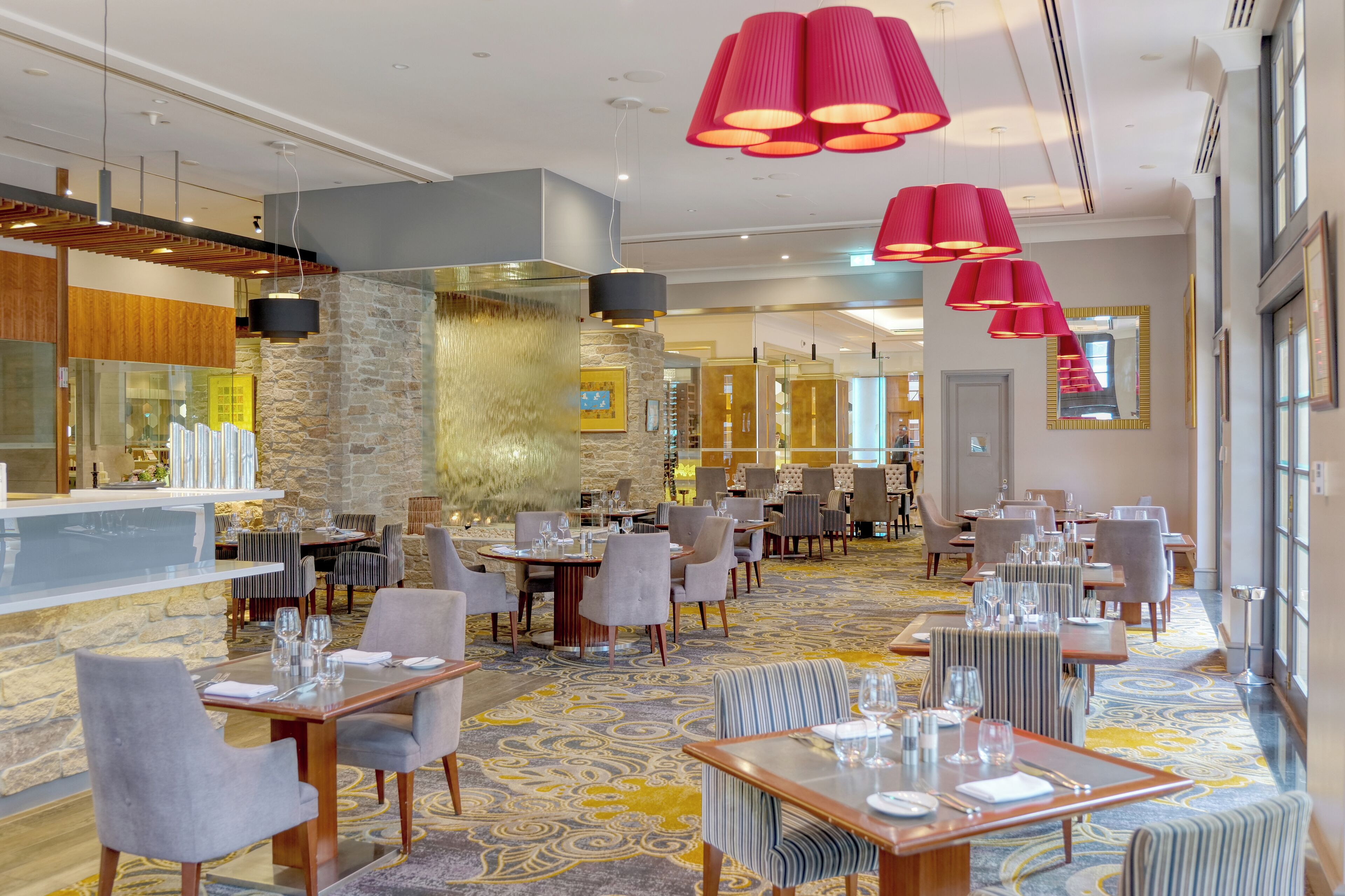 A modern hotel lobby with a stone wall, a bar area, and multiple dining tables with chairs.