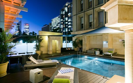 A rooftop pool area with lounge chairs, a small table, and a seating area with a view of the city skyline at dusk.