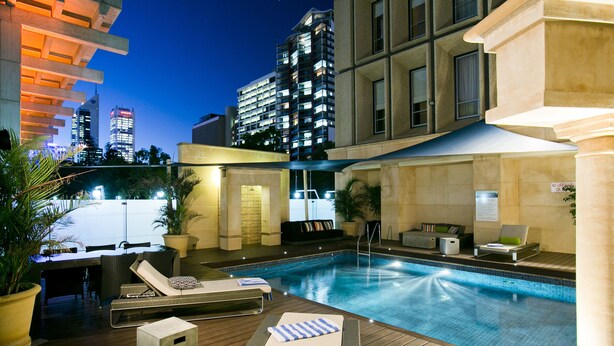 A rooftop pool area with lounge chairs, a small table, and a seating area with a view of the city skyline at dusk.