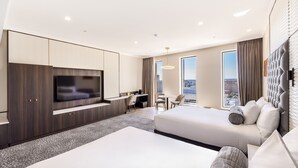 A modern hotel room with a large flat-screen TV, a desk with a chair, two beds, and a city view through large windows.