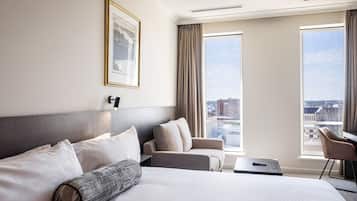 A hotel room with a large bed, a sofa, a small table, and a chair. There is a window with a view of the city.