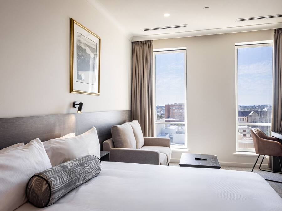 Ein Hotelzimmer mit einem großen Bett, einer Sitzecke, einem Schreibtisch und einem Sessel. Zwei große Fenster bieten Blick auf die Stadt.