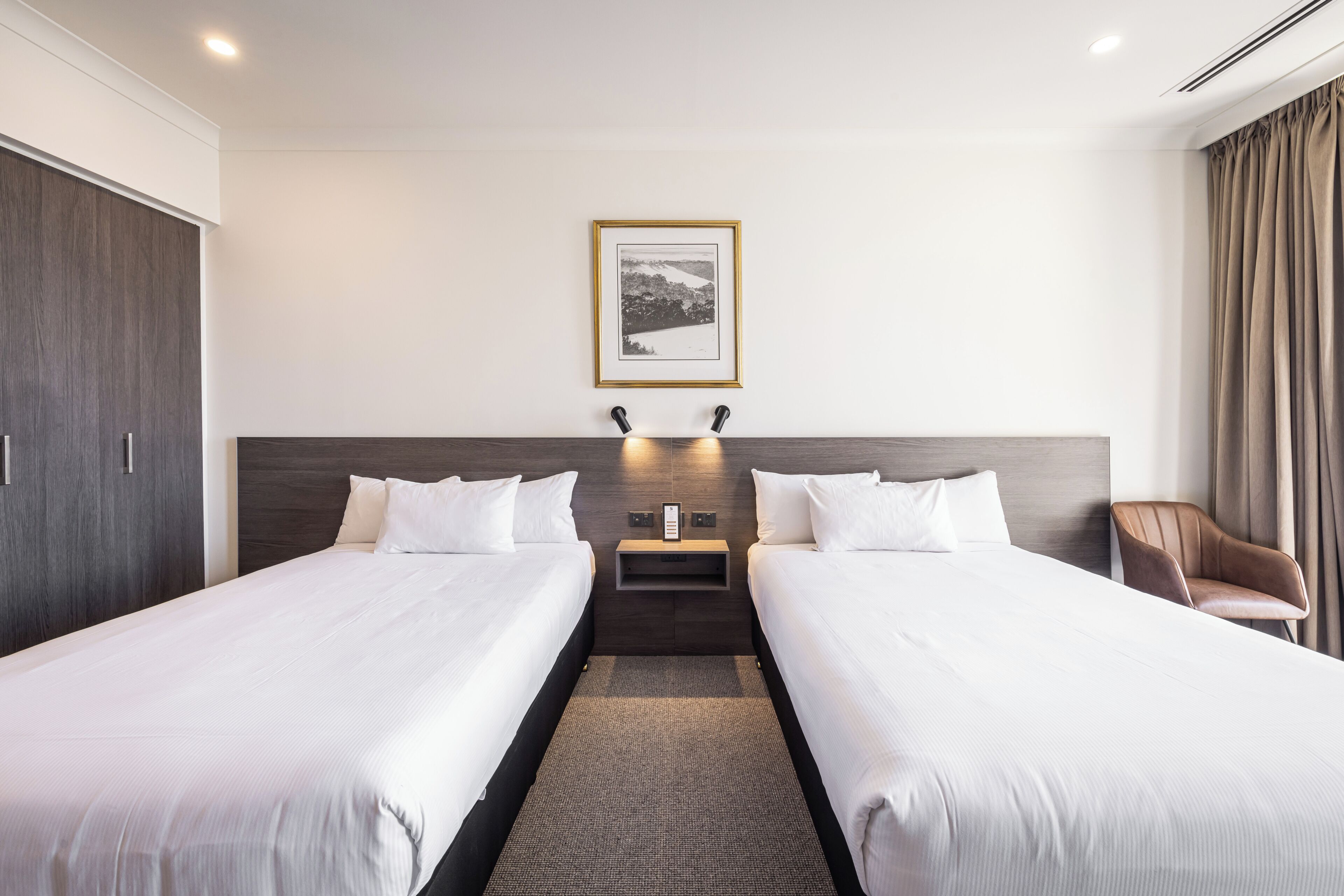 A hotel room with two beds, a nightstand, a framed picture on the wall, and a chair.