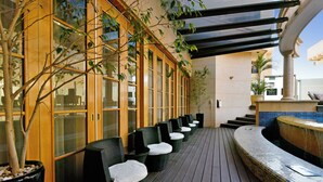 A row of black wicker chairs with white cushions, a tiled bench, and a potted tree on a wooden deck.