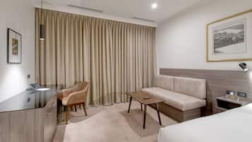 Una habitación de hotel moderna con un escritorio de vidrio, un sofá color beige, una mesita y una cama.