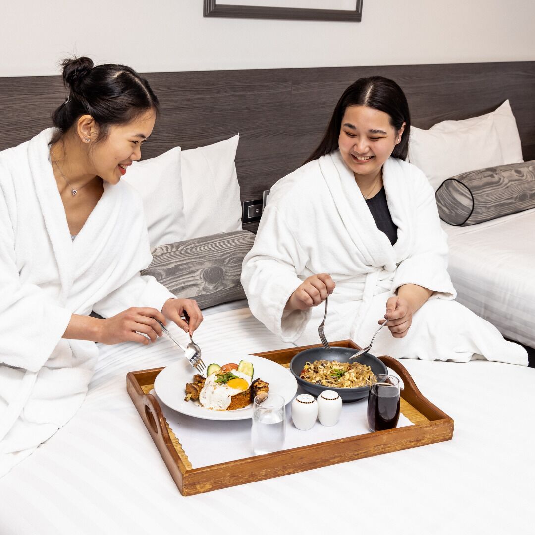 Two individuals in white robes sitting on a bed with a tray of breakfast food, including eggs and noodles, and two salt shakers.