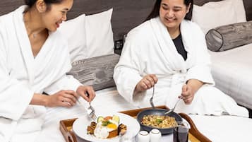 Two individuals in white robes sitting on a bed with a tray of breakfast food, including eggs and noodles, and two salt shakers.