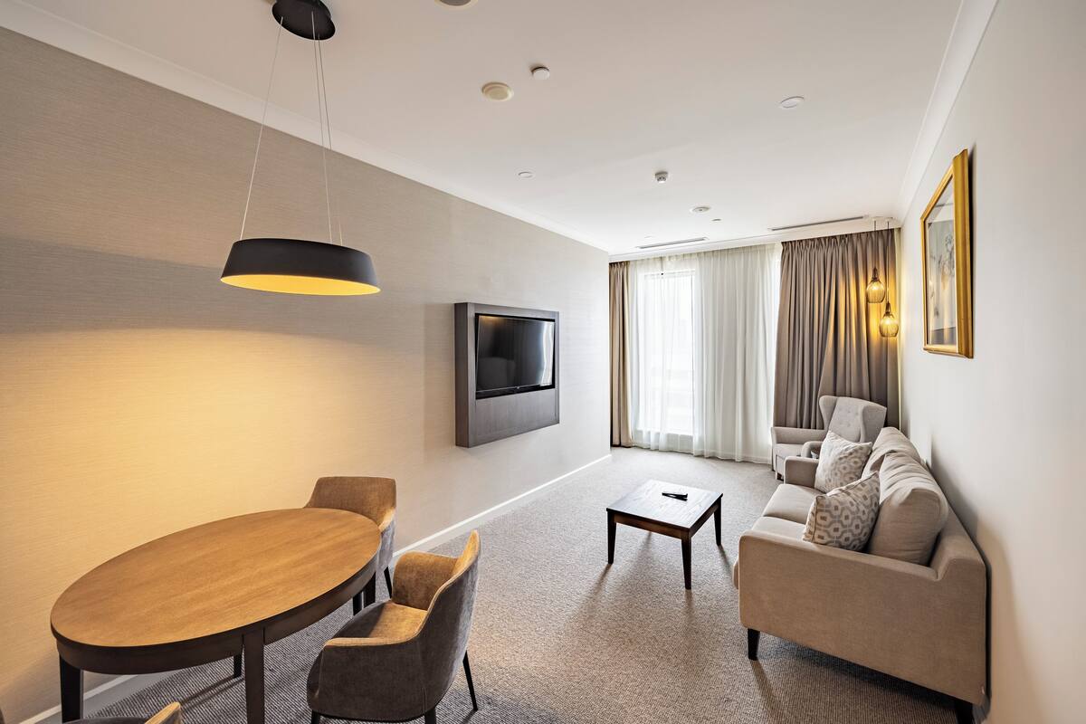 Habitació d'hotel moderna amb taula de menjador, sofà, televisió i un gran finestral amb cortines.