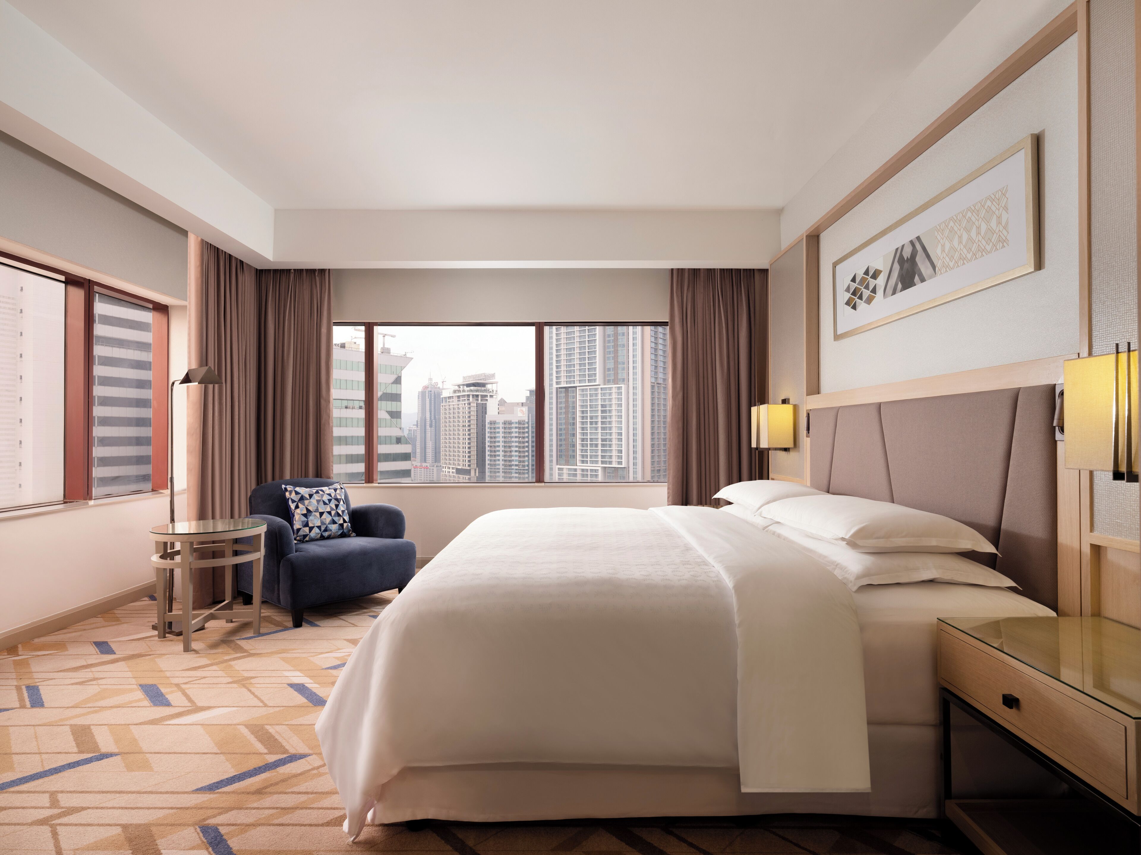 A hotel room with a large bed, a chair, a small table, and a view of the cityscape through the window.
