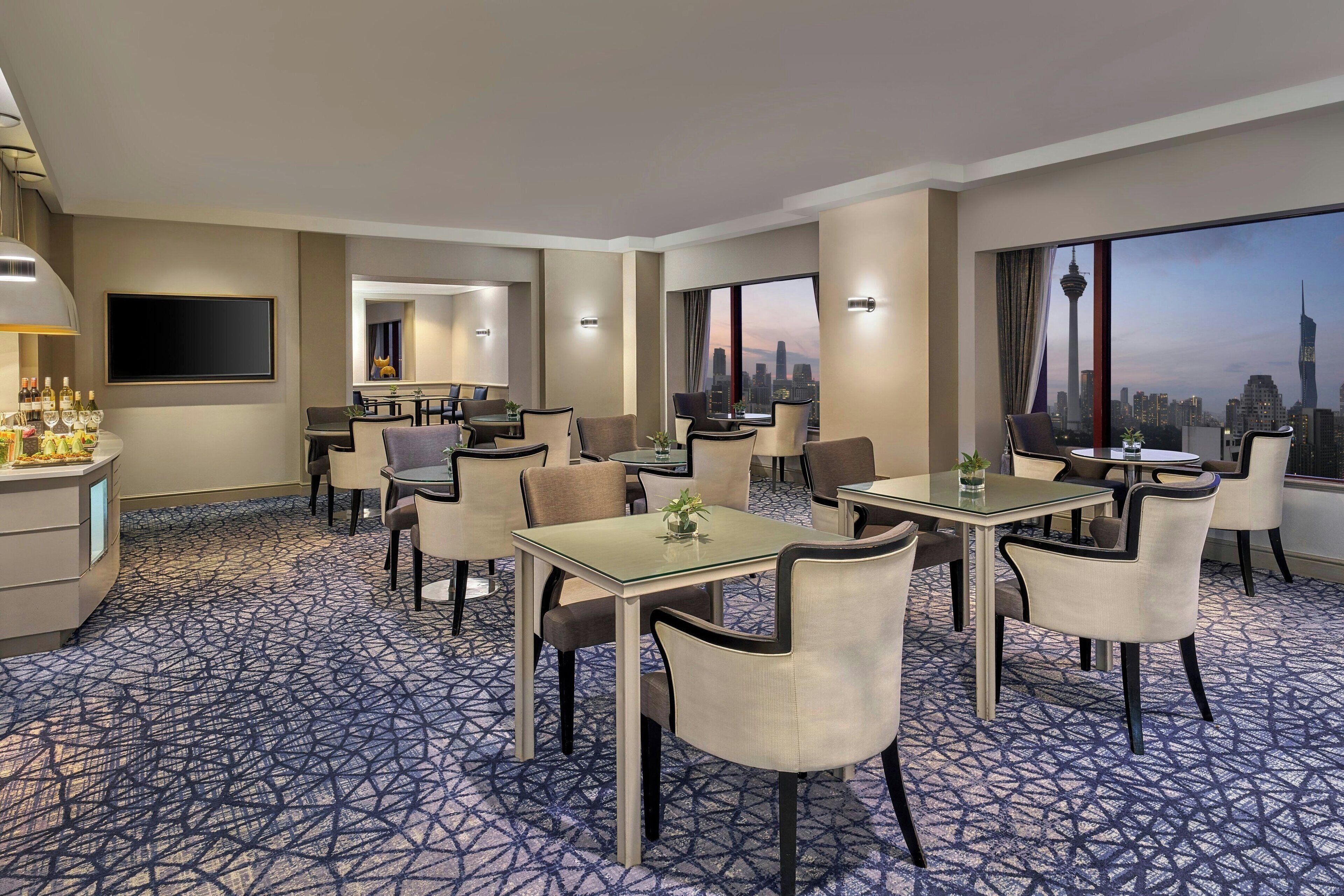 Un comedor de hotel con varias mesas y sillas, una zona de bar y un amplio ventanal con vista al horizonte de la ciudad.