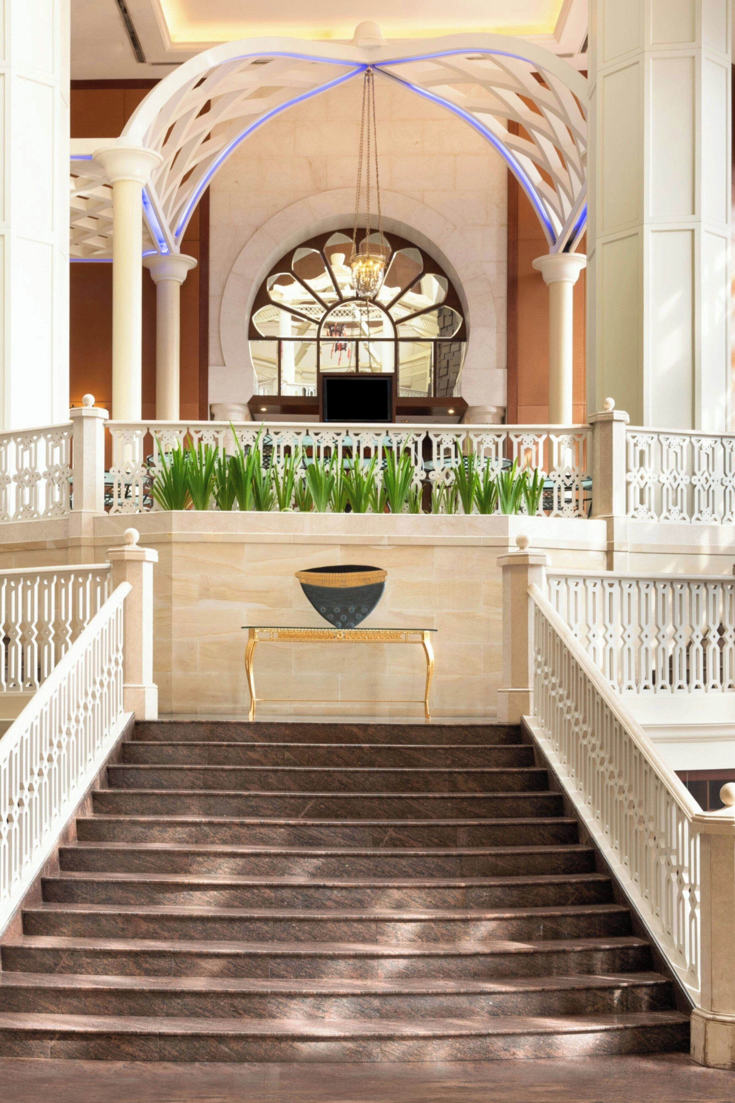 Una majestuosa escalera con suelo de mármol, balaustradas blancas y un arco decorativo con una araña.