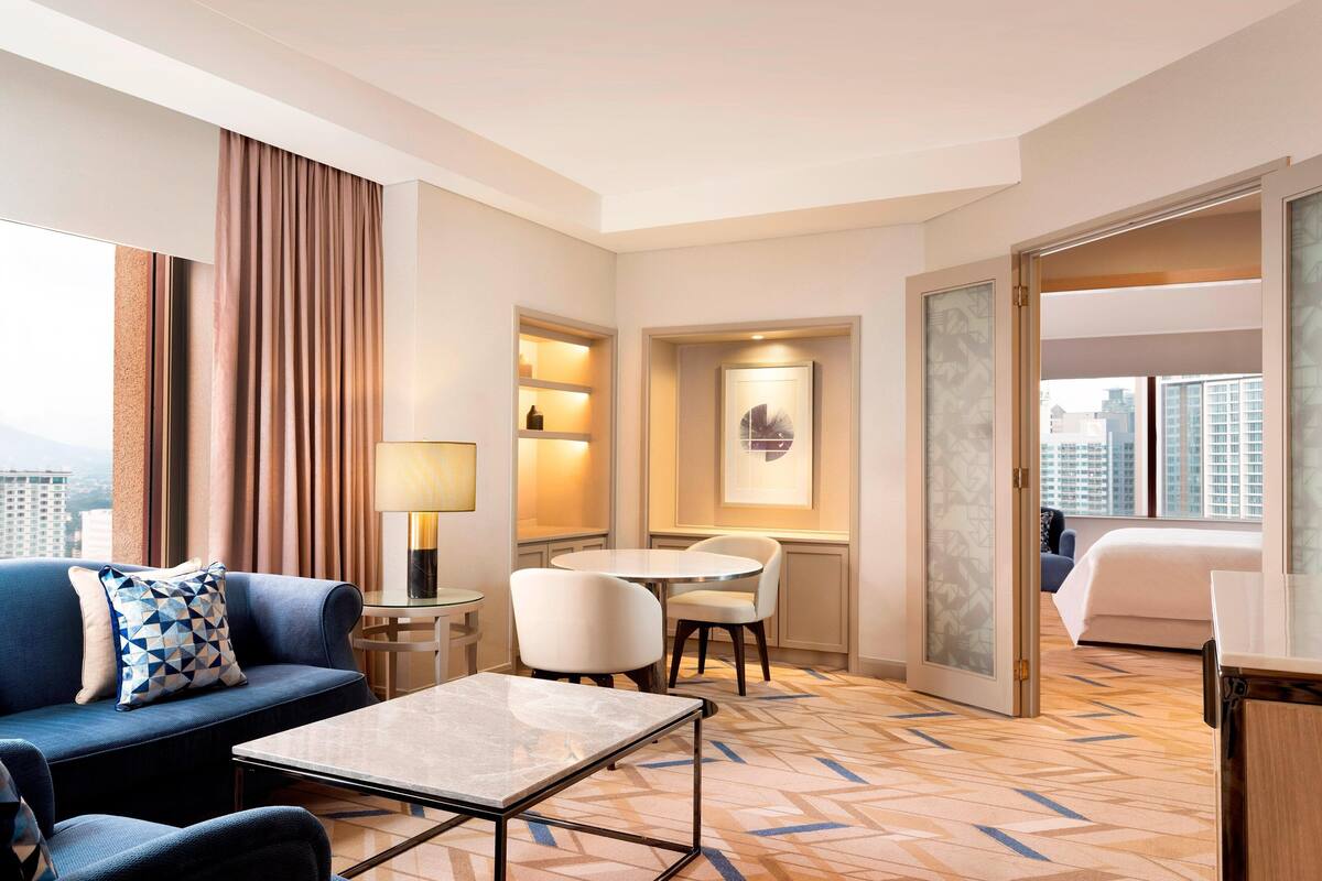 A hotel room with a sofa, a small table, a desk with chairs, a bed, and a city view.
