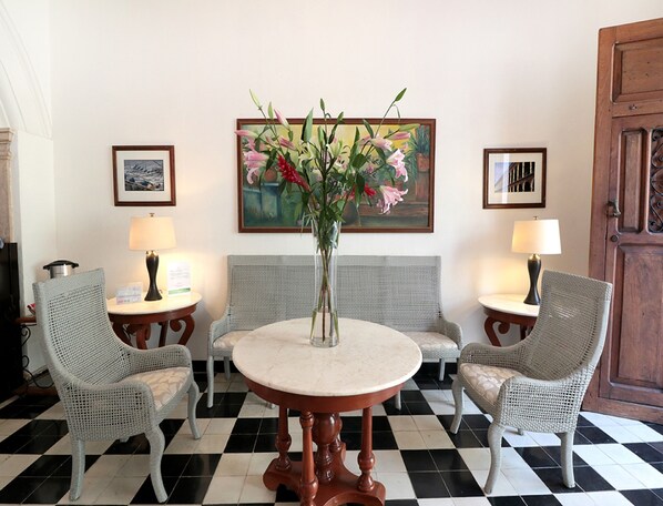 Un hall d’entrée avec un sol à carreaux, deux fauteuils en osier, une table ronde ornée d’un vase de fleurs et deux lampes.