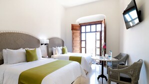 Habitación de hotel con dos camas, un televisor montado en la pared, una mesita con sillas y una ventana con vistas.