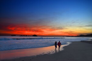 Escena de playa con dos personas caminando por la orilla al atardecer.