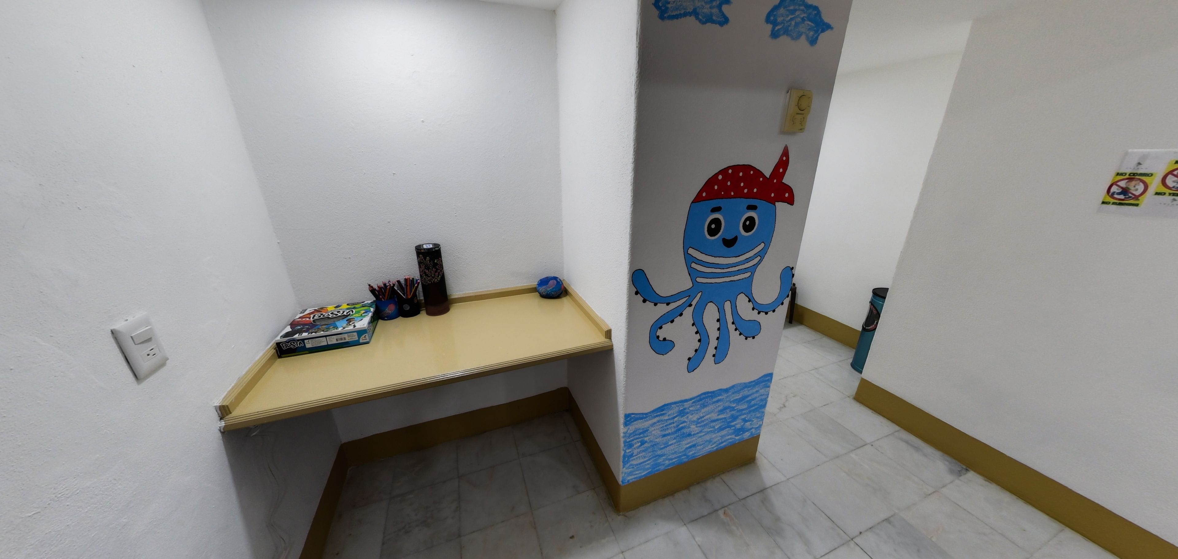 A narrow hallway with a small desk, a wall with a cartoon octopus, and a trash bin.
