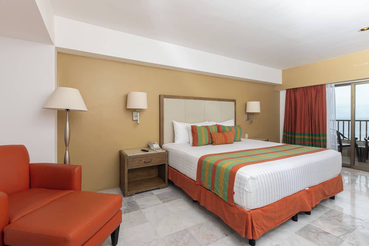 A hotel room with a large bed, a nightstand, a chair, and a balcony with curtains.