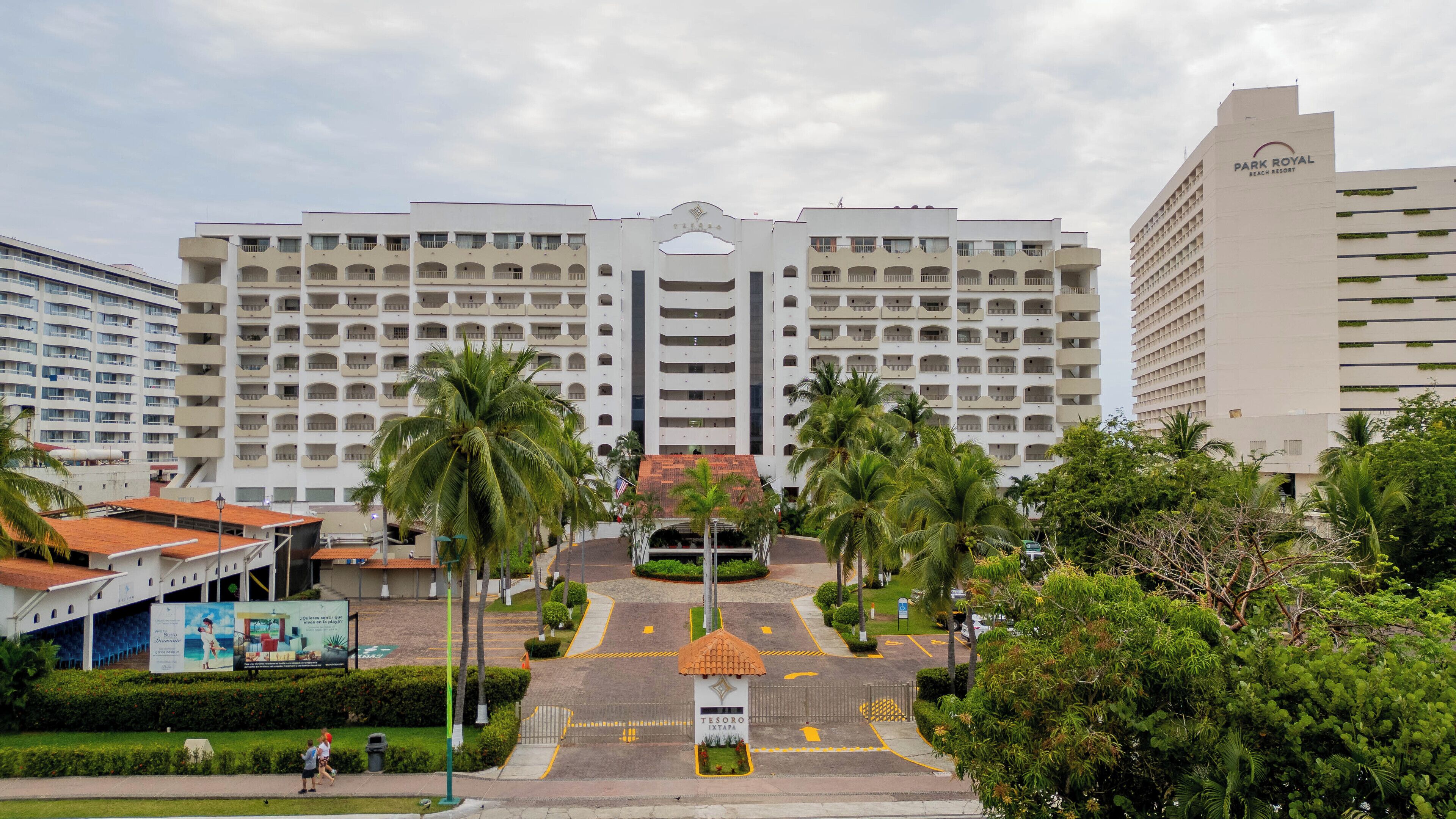 Um hotel grande com vários andares e varandas, uma entrada central com telhado decorativo e palmeiras em primeiro plano.