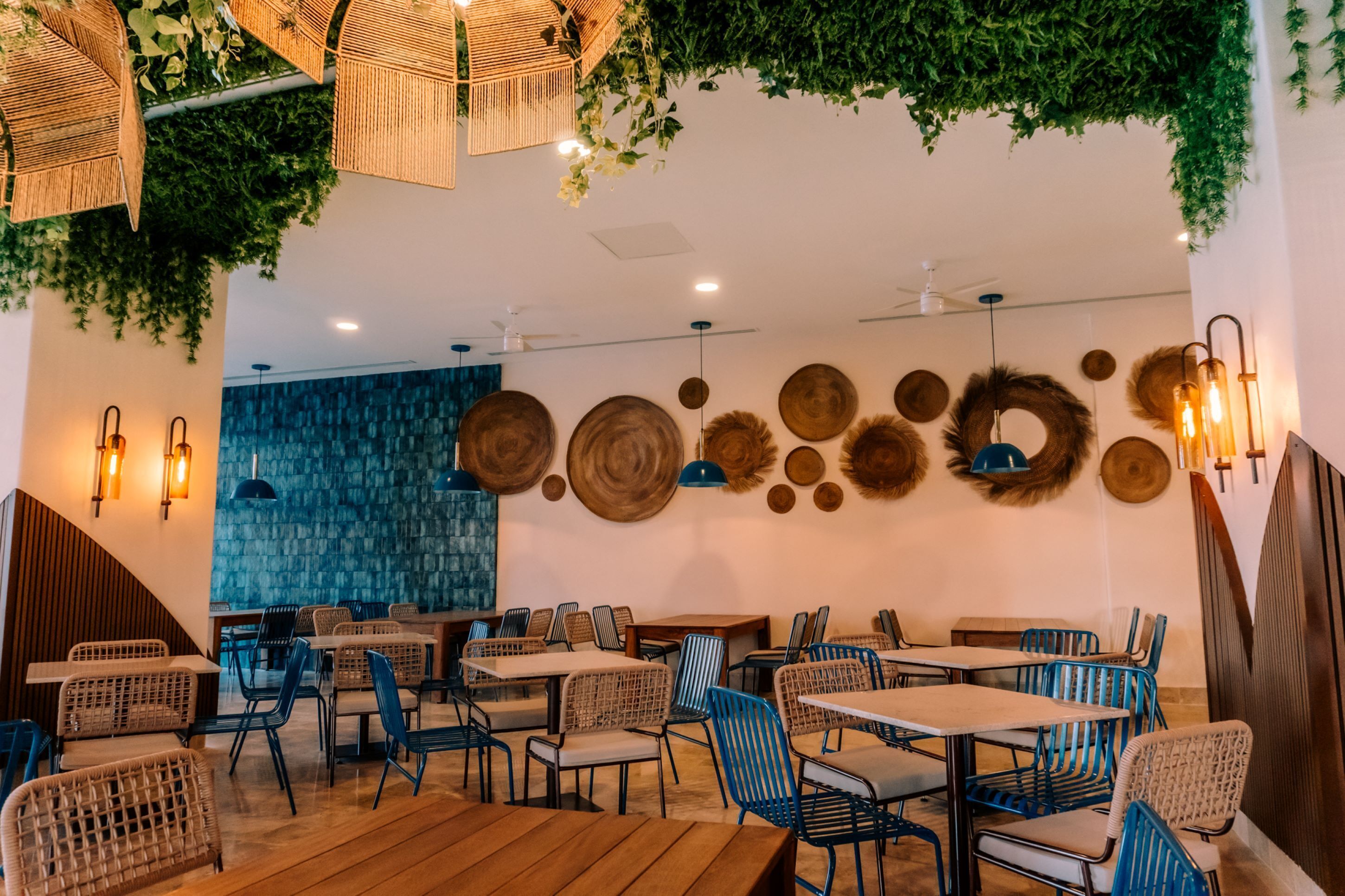 L’intérieur d’un restaurant, avec des tables et des chaises en bois, des plantes suspendues et des œuvres d’art décoratives accrochées aux murs.