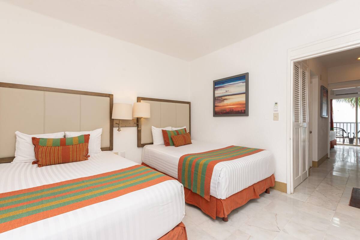 A hotel room with two beds, a painting on the wall, and a view through an open door.